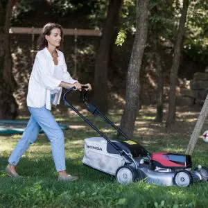 a woman in jeans and a white top pushing a lawnmower past some trees