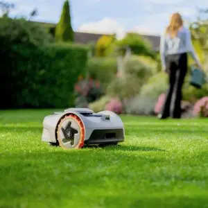 the Segway Navimow i105E Robot Mower cutting a lawn, with a woman watering plants in the background