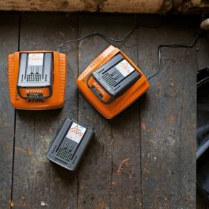 3 garden machinery batteries, 2 of which are in chargers, all on top of a wooden table