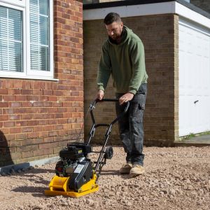 a man using a Handy THLC29140 Petrol Plate Compactor outside a house, with a garage in the background