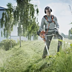 a man wearing ear defenders, gloves and protective boots, stood on a grassy slope, using a brush cutter