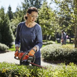 a woman wearing goggles and gloves, operating a Husqvarna hedge trimmer