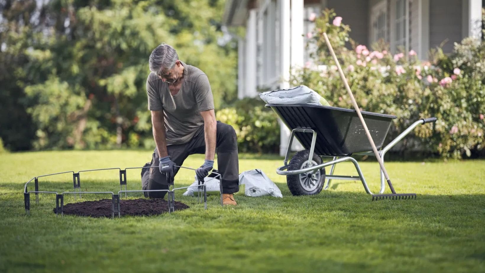 a professional gardener fixing a metal ring around a patch of soil on a lawn, with a wheelbarrow and garden tools in the background