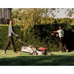 a woman mowing a lawn, with a man using a hedge trimmer in the background