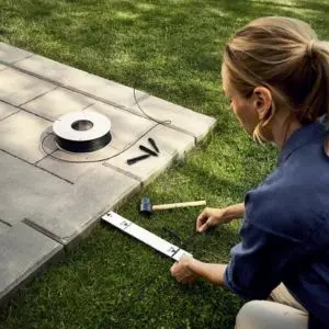 a woman with a ruler, mallet, pegs and wire, installing a robotic mower's boundary wire next to a patio