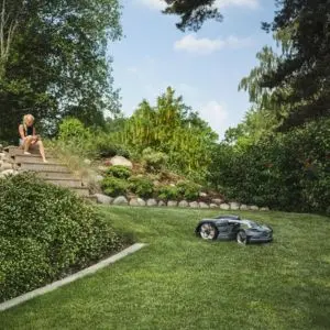 a robot mower moving down a gentle slope, with a woman sat on steps, using a mobile device, in the background