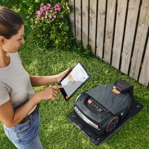 a woman using an app on a tablet, stood next to a robot mower on its charging station