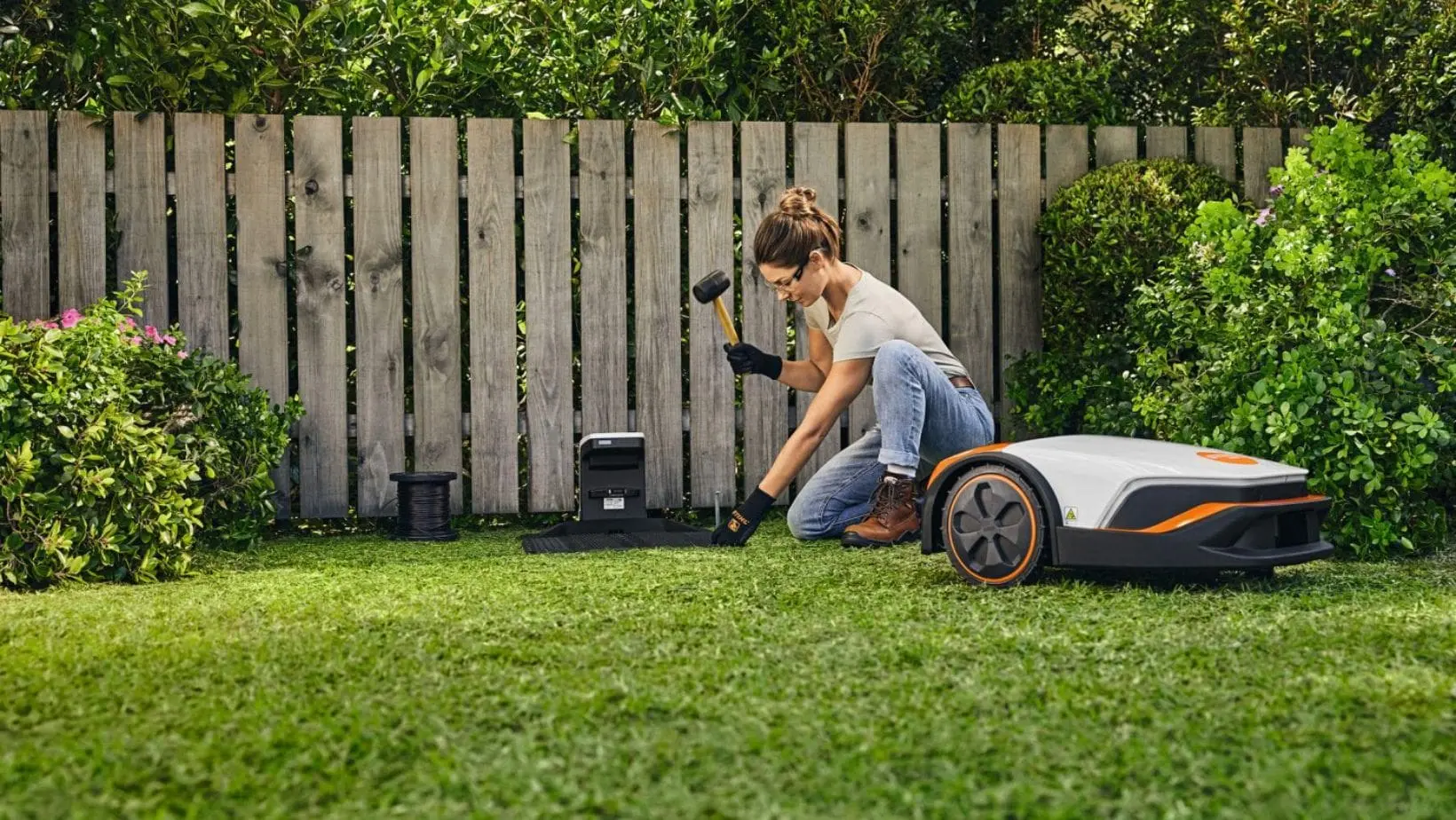 iMow installation-min a woman installing a robotic mower charging station on a lawn, with a white robot mower next to her and a picket fence and shrubs in the background