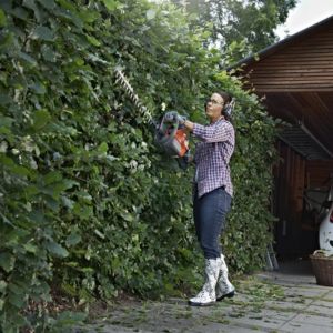 a woman wearing ear defenders and gloves, trimming the side of a tall hedge with a petrol hedge cutter