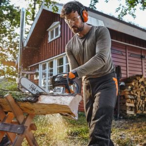 a man wearing ear defenders and safety glasses, stood outside a wooden house, using a petrol chainsaw to cut a log
