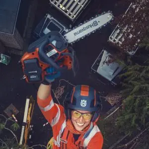 a woman in Husqvarna-branded PPE, smiling and holding aloft a Husqvarna top handle chainsaw