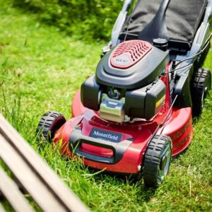 a close up of a red Mountfield petrol lawnmower cutting grass in front of a slatted wooden bench