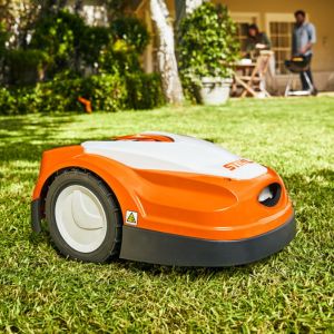 the STIHL iMow Robotic Mower on a lawn, with a man, house, trees and plants in the background