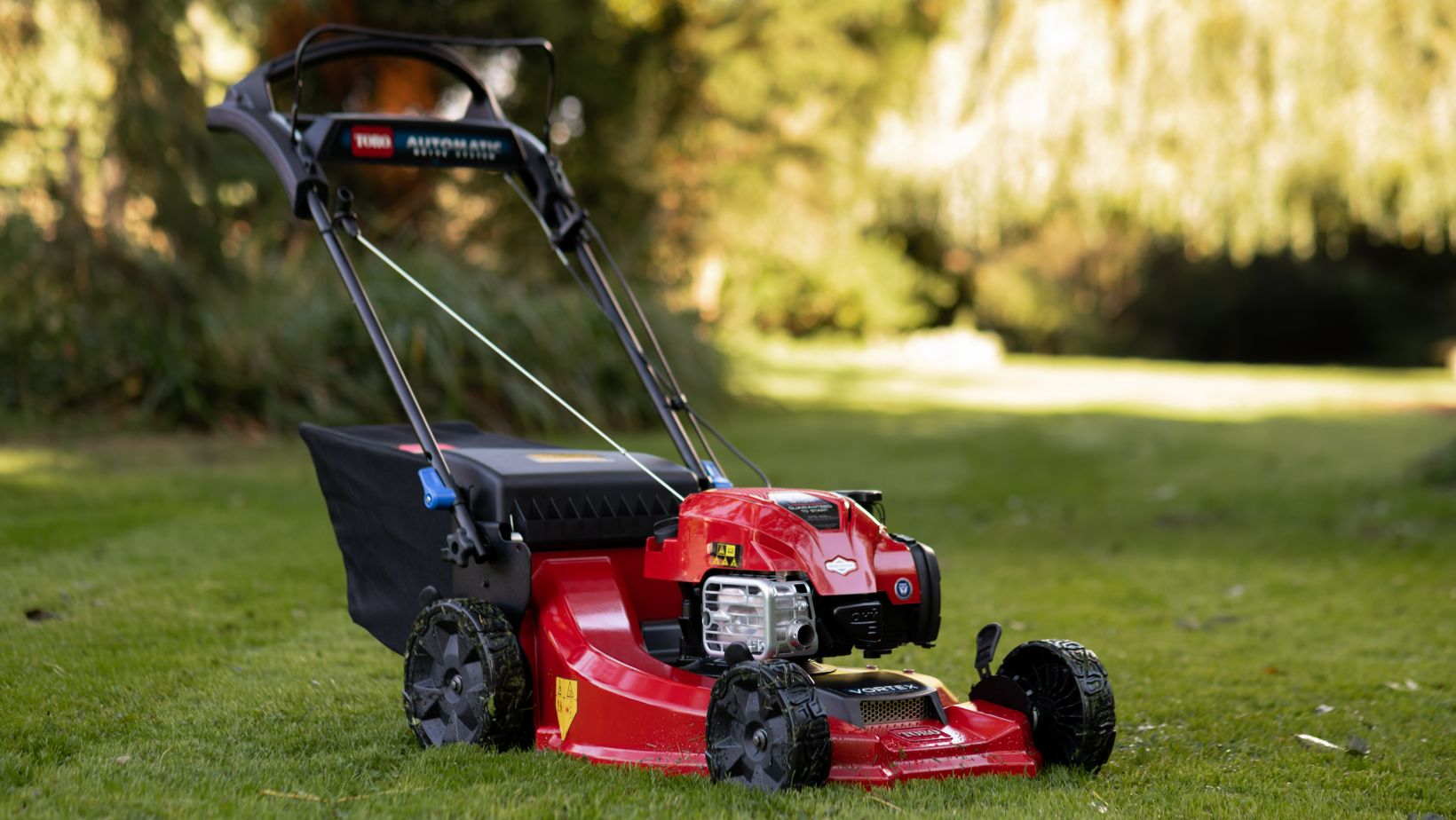 Toro Super Recycler-min a red Toro Super Recycler Petrol Lawnmower sat on a lawn with trees and shrubs in the background