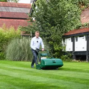 a man using the Allett Buckingham 24H Cylinder Lawnmower to create lawn stripes, with a large house to the right and rear