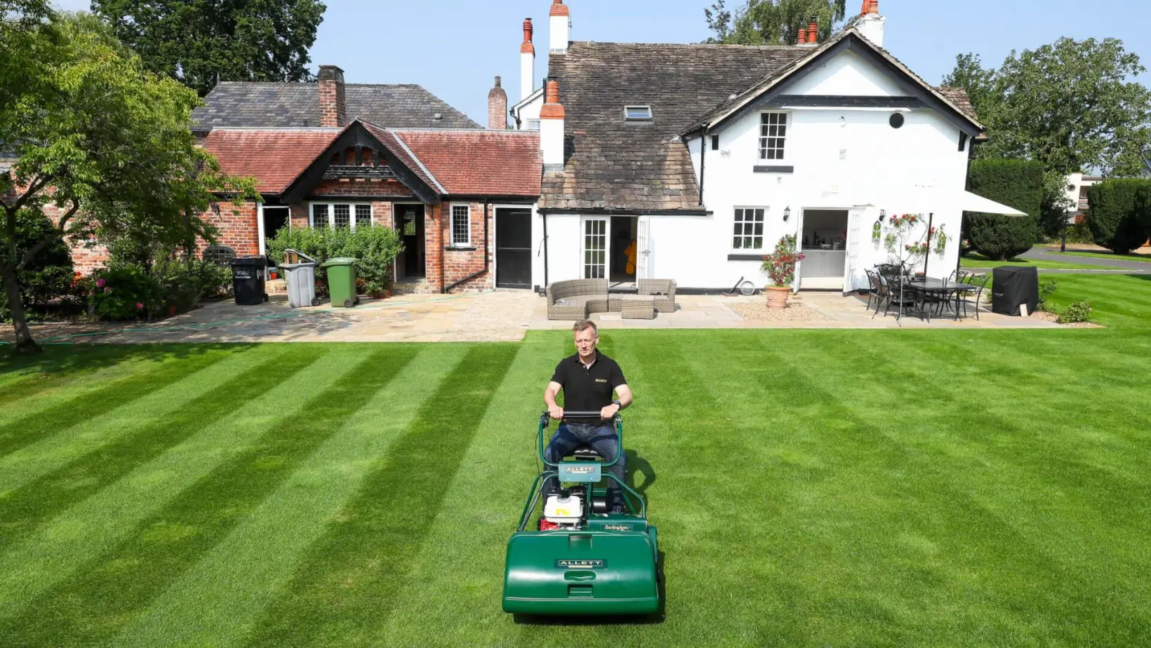 a man stood behind an Allett Buckingham Cylinder Lawnmower, on a striped lawn, in front of a large house