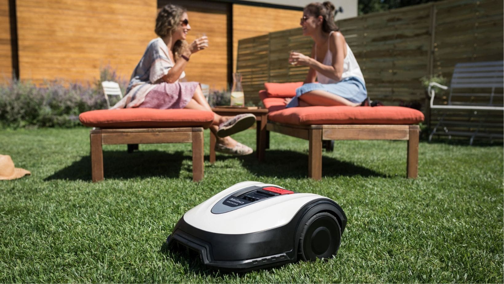 a Honda Miimo robotic mower in front of 2 women, who are sat on cushioned wooden seating in the garden