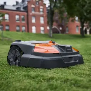 an orange and black Husqvarna Automower on a lawn, outside a red-brick stately home