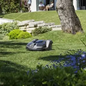 a Husqvarna Automower travelling down a sloping lawn, with a tree, shrubs, seated couple, and house in the background