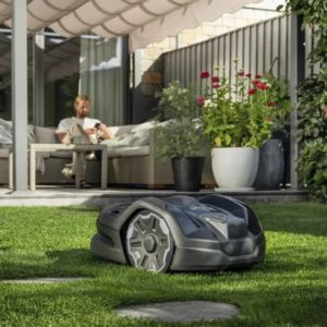 the Husqvarna Automower® 305E NERA mowing a lawn, with a man sat on garden furniture in the background