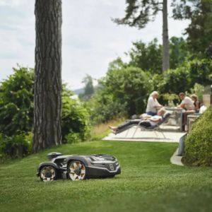 the Husqvarna Automower® 435X AWD Robot Mower travelling across an undulating lawn, with trees, shrubs, and people relaxing on garden furniture in the background