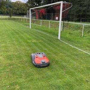a Husqvarna robot mower on a basic football pitch, with a wood in the background