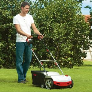 a man wearing jeans and a white t-shirt, pushing the AL-KO 38 E Electric Scarifier across a lawn, with trees in the background
