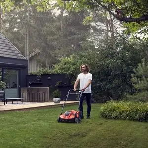 a man pushing a Husqvarna aerator across a lawn, with trees, shrubs, decking and a house in the background