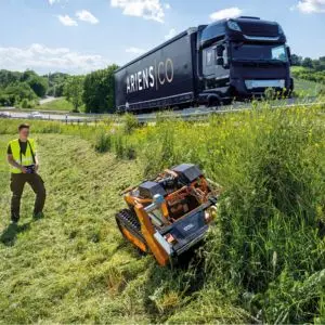 a man wearing a high-visibility jacket, using a remote control to operate the AS-Motor AS 1000 OVIS RC Petrol Flail Mower on a steep roadside verge, with an Ariens articulated lorry on the road in the background