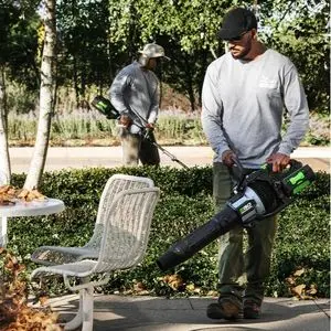 a man wearing a flat cap, using the EGO LBX1000 Cordless Blower to clear leaves in a park