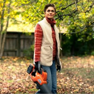a woman with short hair, wearing a gillet, holding the Husqvarna 230iB Leaf Blower, and surrounded by fallen leaves in an autumn garden