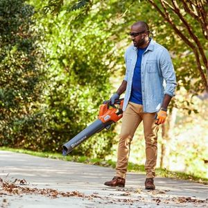 a man dressed in a blue shirt and beige trousers, using a Husqvarna 230iB Cordless Blower to clear leaves from a driveway