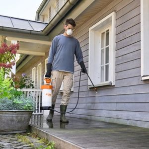 a man wearing a protective mask, carrying the STIHL SGA 30 Cordless Sprayer across a decked area, outside a house