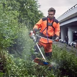 a man dressed in orange and black PPE, using the Husqvarna 535iRXT Brushcutter to cut overgrowth on a verge near a bridge