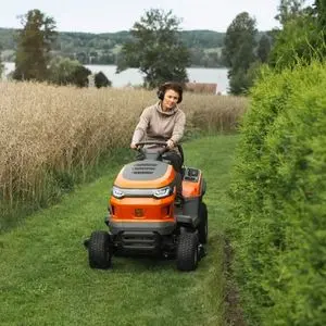 a woman driving the Husqvarna TS 215T Garden Tractor on a lawn, between long grass and hedging, with trees and a river in the background