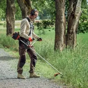 a man wearing ear defenders, using a Husqvarna brushcutter to cut overgrown grass, near trees, at the side of a gravel path