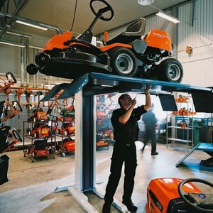 a technician working on the underside of a Husqvarna riding mower, which is suspended above him on a vehicle lift