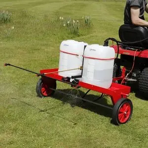a riding mower towing the SCH HGPS Sprayer across a lawn, with a clear liquid being released from its 2 containers