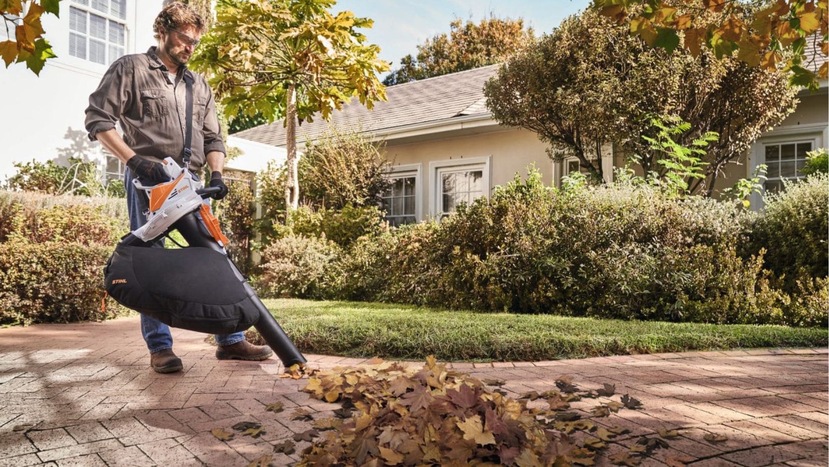 a man on a residential driveway vacuuming up a pile of leaves using the STIHL SHA 56 Cordless Handheld Leaf Blower & Vacuum Shredder