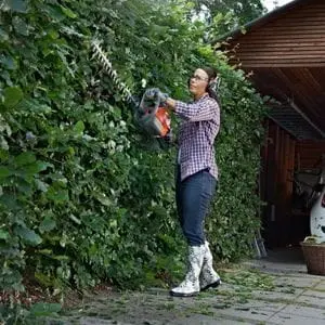 a woman wearing ear defenders and gloves, using the Husqvarna 122HD60 Hedge Cutter to trim a hedge