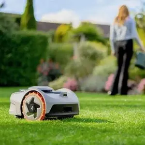 a Segway Navimow i105E Robotic Mower on a lawn, with a woman watering plants in the background