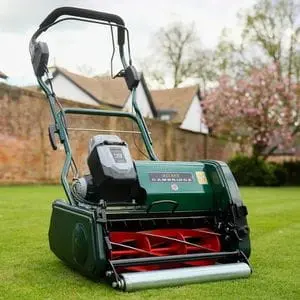 the Allett Cambridge 43 Cordless Cylinder Mower on a perfectly mown lawn, with trees, a wall, and the tops of two white-walled buildings in the background