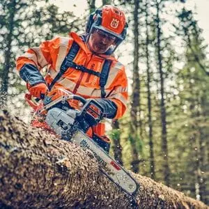 a man wearing orange chainsaw PPE, using a Husqvarna chainsaw to cut a large log