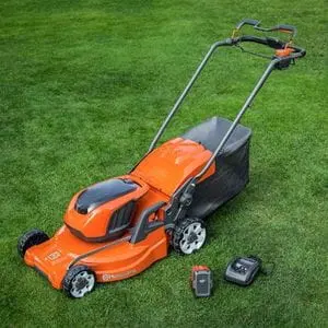 an orange Husqvarna cordless mower, together with a battery and charger, all displayed on a lawn