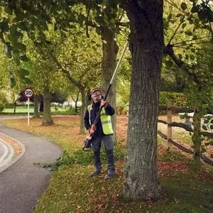 a man wearing a high-visibility jacket, stood on the side of a tree-lined road, using the Husqvarna 530iPT5 Pole Saw to prune a tree, with a 30mph speed limit sign in the background