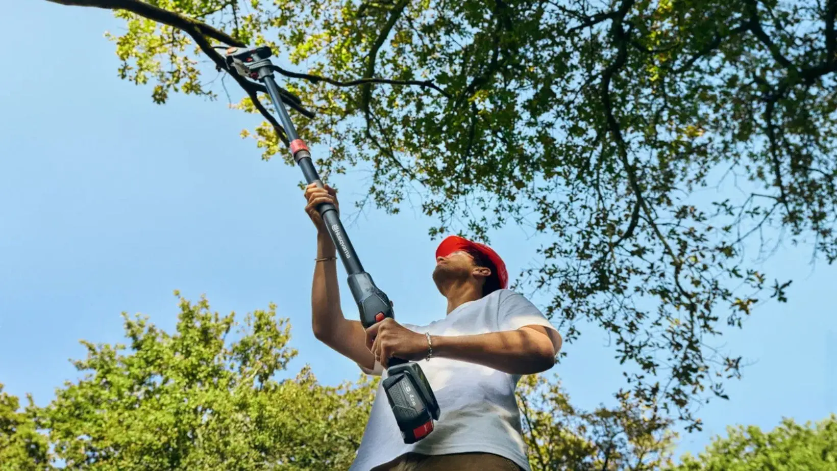 Husqvarna Aspire™ PE5-P4A (1) a clear day, with a man wearing an orange hard hat, using the Husqvarna Aspire™ PE5-P4A Pole Pruner to prune overhanging branches on a tree