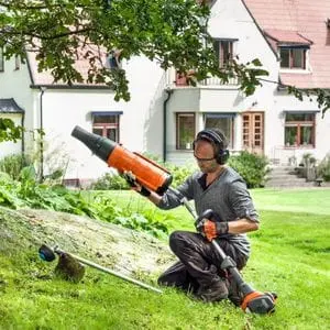 a man sat on a lawn, fitting a leaf blower attachment to a Husqvarna combi tool, with a tree and large white house in the background