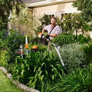 a man using the STIHL KMA 80 R Cordless KombiMotor with hedge trimmer attachment to prune shrubs in a garden, with a wheelbarrow full of other multi attachments next to him