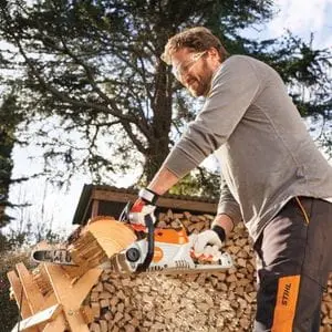 a man using the STIHL MSA 70 Cordless Chainsaw to cut a log on a sawhorse, with a full logstore in the background
