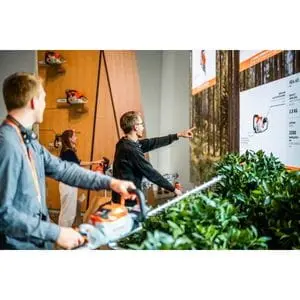 a man in an office, holding a STIHL hedge trimmer, while another man points at a board to explain the tool's capabilities, while a woman looks on